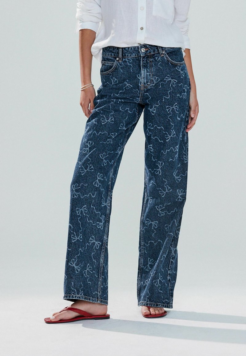 Jeans din denim închise la culoare, cu o croială lejeră, având modele abstracte de fundițe albe, design cu cinci buzunare și cusături vizibile. Purtați cu sandale roșii.