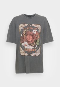 Grå, oversized t-skjorte laget av bomull. Forsiden har et fargerikt trykk med en sol, blomster og sommerfugler, rammet inn av dekorative elementer og tekst.