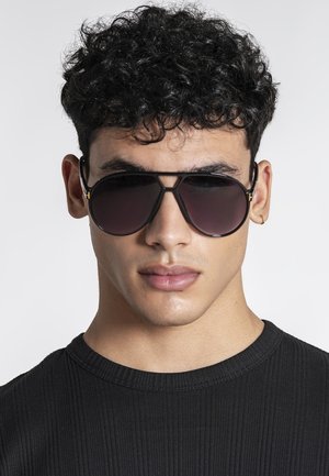 Junger Mann mit lockigem Haar, der schwarze Piloten-Sonnenbrillen und ein schwarzes, geripptes Shirt mit Rundhalsausschnitt trägt, auf einfachem Hintergrund frontal blickend.