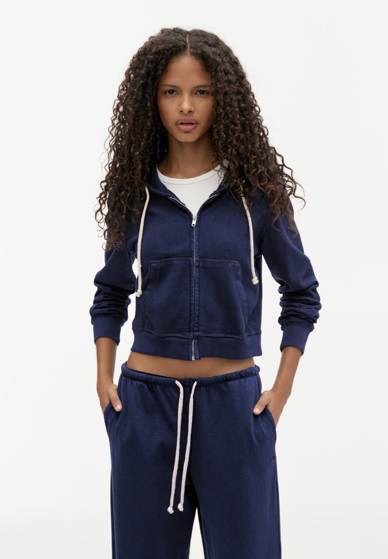 Felpa cropped blu navy con zip, realizzata in tessuto morbido, con tasca frontale e cappuccio con cordoncino. Abbinata a pantaloni della tuta blu navy.