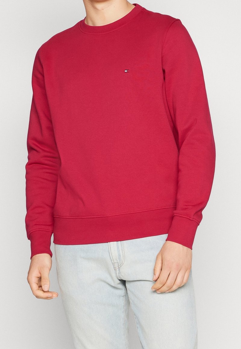 Homme portant un sweat-shirt rouge uni à manches longues et un jean bleu clair sur un fond neutre, les mains détendues le long du corps.