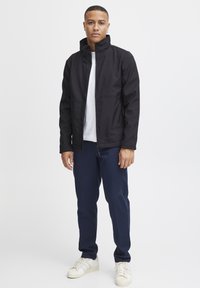 Zwarte softshell jas met een hoge kraag, rits aan de voorkant en twee zijzakken, gecombineerd met marineblauwe broek en witte sneakers.