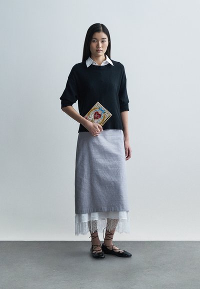 Fekete kötött pulóver, alatta fehér galléros ing, szürke szoknya csipkés szegéllyel, fekete fűzős lapos cipő, dekoratív könyvet tartva.