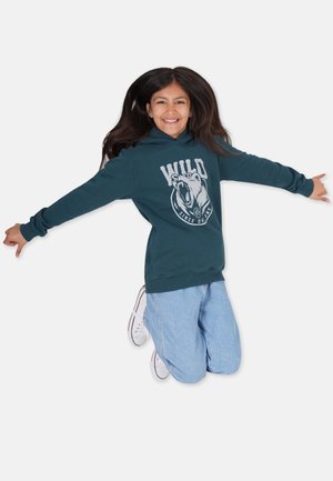 Mørkegrøn hættejakke med et stort bjørne-grafik og "WILD" tekst. Kombineret med lyseblå jeans og hvide sneakers. Barn der hopper med udstrakte arme.