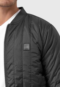 Black quilted bomber jacket with a zipper, ribbed collar, and a square black logo patch on the left chest. Smooth, lightweight material.