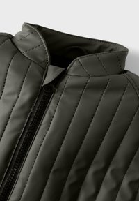 Dunkelgrüne gesteppte Jacke mit diagonaler Naht und einem schwarzen Reißverschluss, teilweise am Kragen zugezippt vor einem schlichten Hintergrund.