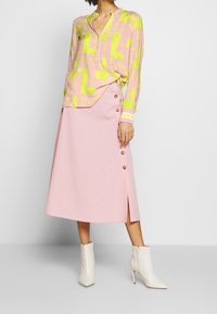Blusa de manga larga con un patrón en rosa y amarillo, con escote en V, combinada con una falda rosa que presenta detalles de botones en el lado y una abertura en la parte delantera. Botas blancas hasta el tobillo.