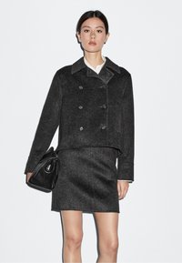 Giacca nera in misto lana a doppio petto con colletto a punte, abbinata a una gonna fitted dello stesso tessuto. La modella tiene una borsa nera.