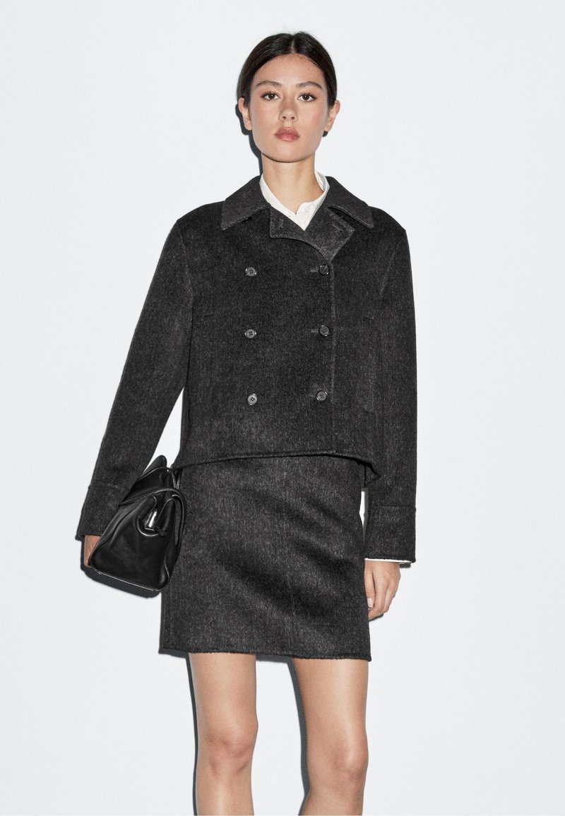 Giacca nera in misto lana a doppio petto con colletto a punte, abbinata a una gonna fitted dello stesso tessuto. La modella tiene una borsa nera.