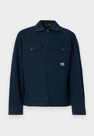 Marineblå jakke med glidelås foran, to brystlommer med knapper og en liten rektangulær lapp på venstre lomme.