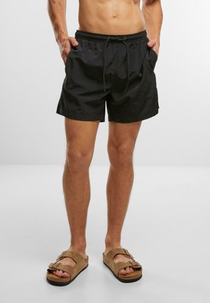 Schwarze Schwimmshorts mit einem elastischen Bund, verstellbarem Kordelzug und Seitentaschen. Tragen mit braunen Slippern.