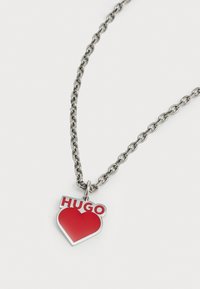 Metallkjede med en rød hjerteformet anheng som har ordet "HUGO" i fete, hvite bokstaver på en sølvfarget bakgrunn.