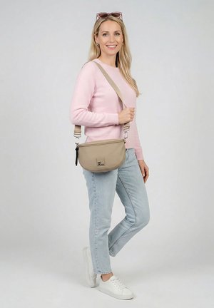 Lächelnde Frau in pinkem Pullover, hellblauer Jeans, weißen Turnschuhen, Sonnenbrille auf dem Kopf, die eine beigetönte Umhängetasche mit verstellbarem Riemen vor schlichtem Hintergrund hält.