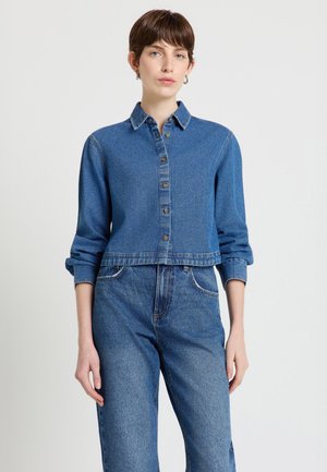 Donna con capelli castani corti che indossa una camicia di jeans blu abbottonata e jeans di jeans blu a vita alta su uno sfondo chiaro e semplice.