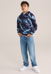 Temno modra kapuca z modrimi in teal vijugami, kombinirana z svetlo modrimi kavbojkami in belimi supergami. Kapuca ima sprednji kenguru žep.