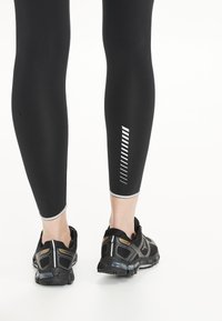 Zwarte leggings met reflecterende accenten bij de enkels, aangesloten ontwerp, gecombineerd met zwarte sportschoenen met een textuur en grijze details.