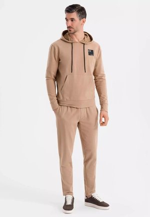 Sudadera beige con bolsillo frontal, cordón y parche con logo, combinada con pantalones de chándal ajustados a juego y zapatillas marrones oscuras. Textura suave.