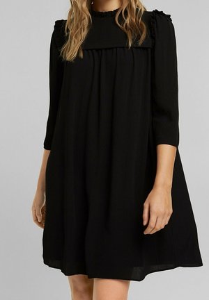 Frau trägt ein locker sitzendes schwarzes Kleid mit dreiviertel Ärmeln und gerafftem hohem Kragen vor einem einfarbig hellgrauen Hintergrund.