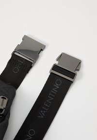 Les sangles en nylon noir avec des boucles métalliques, arborant un logo. Surface texturée sur une boucle, surface lisse sur l'autre.