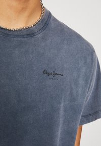 Camiseta de algodón azul marino de manga corta, con un sutil logo en negro de "Pepe Jeans London" en el área del pecho izquierdo. Textura suave.