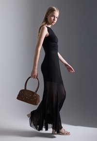 Jeune femme marchant dans une robe sans manches noire avec des panneaux de jupe transparents, portant un sac à main marron avec de petites clous, chaussée de sandales à talons blancs.