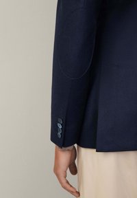 Blazer di lana blu navy con maniche dotate di toppe sui gomiti, caratterizzato da quattro bottoni scuri e un fit sartoriale, che si restringe dolcemente verso il polso.