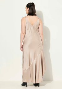 Beige Satin-Maxi-Kleid mit V-Ausschnitt und asymmetrischem Saum. Besitzt dünne Träger und eine glatte Textur. Schwarze Knöchelstiefel runden das Outfit ab.