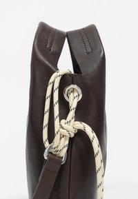 Ciemnobrązowa skórzana torba z białym sznurkiem ściągającym przeprowadzonym przez metalowe oczko, z naszytymi uchwytami i skórzanym paskiem przymocowanym do metalowego pierścienia.