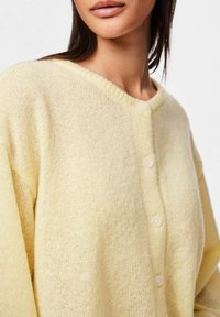 Rumena pletena jopica z okroglo izreza, s tremi gumbi spredaj. Mehka tekstura in ohlapen kroj poudarjata udoben dizajn.