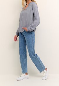 Blouse gris clair avec un col en V, manches longues et poignets froncés, associée à un jean bleu droit et des baskets blanches.