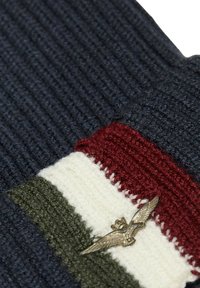 Berretto lavorato a maglia in navy, bordeaux e crema con una striscia verde. Presenta un emblema in metallo a forma di aquila e un motivo a coste testurizzate.