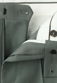 Pantalon en tissu gris avec une ceinture intérieure rayée blanche et grise, présentant des fermetures par boutons et agrafes.