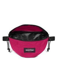 Bolsa de tela redonda color rosa con cierre de cremallera negro y un interior negro, que presenta un parche con el logo. Incluye una correa desmontable para facilitar el transporte.