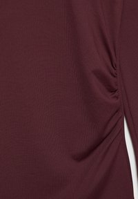 Bordó szövet finom bordázott textúrával, a jobb oldalon gyűrt részletekkel.