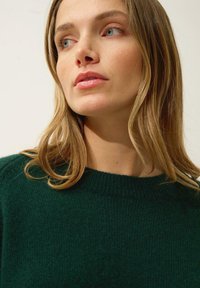 Ciemnozielony, dzianinowy sweter z okrągłym dekoltem i ściągaczami przy mankietach. Miękka tekstura z subtelnymi wariacjami w odcieniu.