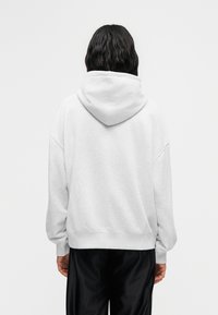 Άτομο με μακριά σκούρα μαλλιά φοράει άνετο λευκό φούτερ με κουκούλα και μαύρο παντελόνι, στέκεται με την πλάτη στραμμένη σε απλό λευκό φόντο.