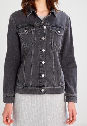 Femme portant une veste en jean noire cintrée avec des boutons argentés sur une robe côtelée gris clair, debout devant un fond uni.