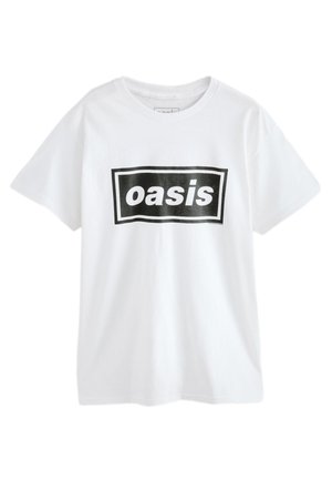 Donna con i capelli lunghi e scuri che indossa una maglietta bianca oversize con il testo "oasis" e pantaloni scuri, in piedi contro una parete chiara e semplice.