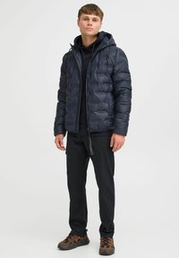 Mørkeblå puffjakke med hætte, udstyret med en quiltet design og lynlås, båret over en sort sweatshirt og sammen med mørke bukser og brune sneakers.