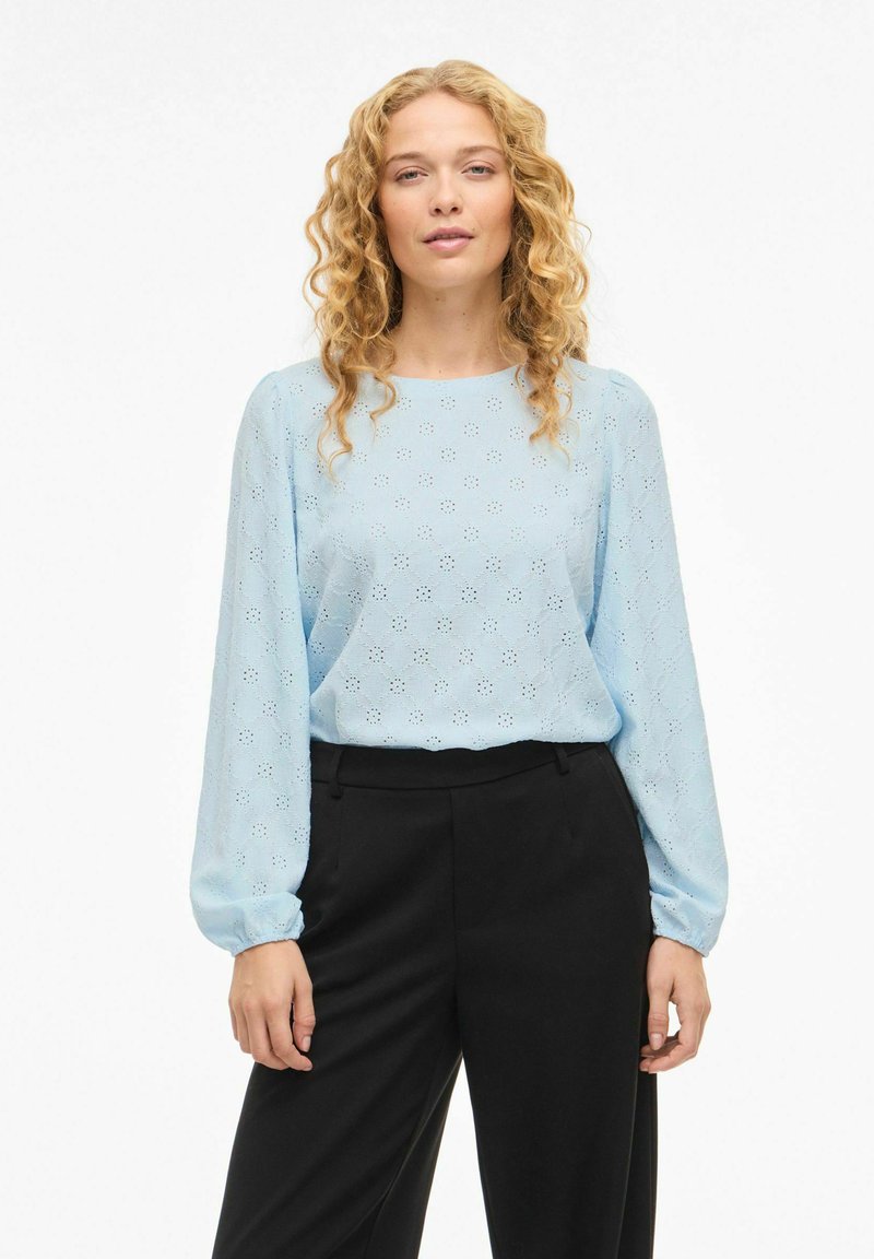 Lyseblå bluse med lange ermer og hullmønster, rund hals og lett rynkede mansjetter, kombinert med svarte bukser. Glatt stofftekstur.