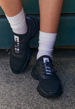 Jambes portant des chaussettes blanches et des baskets noires à lacets avec tissu en mesh et étiquettes de marque, debout sur un béton rugueux près des marches vertes.