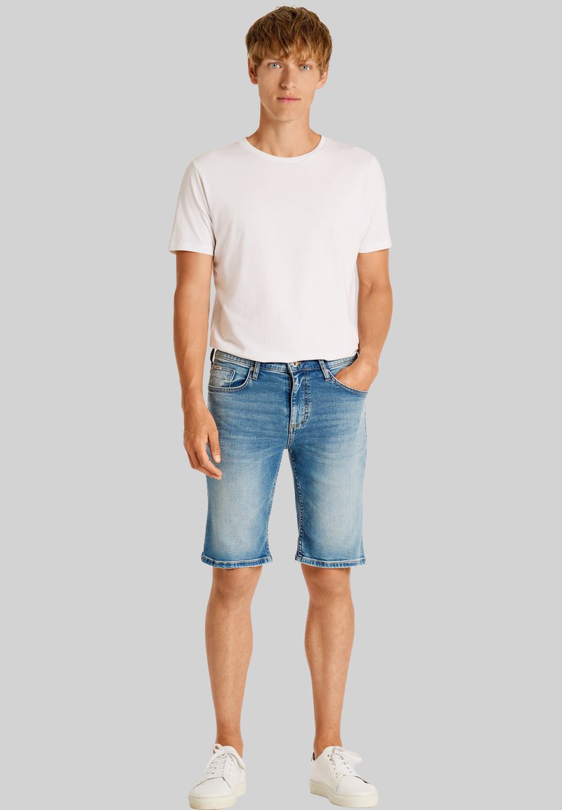 Lättviktig vit bomullströja i kombination med blå denimshorts i knähöjd med uppvikta muddar, bärs med vita sneakers.