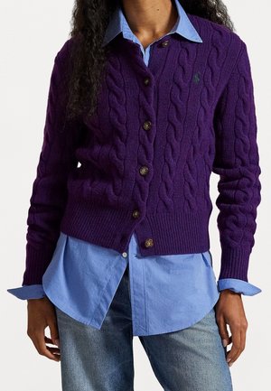 Kobieta ubrana w fioletowy kardigan z grubym splotem i guzikami, nałożony na jasnoniebieską koszulę z kołnierzykiem oraz niebieskie dżinsy, z rękami swobodnie opuszczonymi wzdłuż ciała.