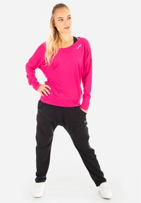 Maglietta rosa a maniche lunghe con scollatura a barca, abbinata a pantaloni neri larghi con tasche. Sneakers bianche completano l'outfit.