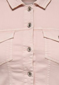 Gros plan sur une veste en jean rose clair montrant des boutons-pression argentés, le col et deux poches à rabat sur la poitrine avec des détails de couture.