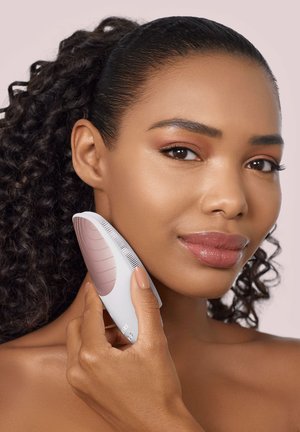 Woman with curly hair holding a white and rose gold facial cleansing brush to her jawline against a light background.