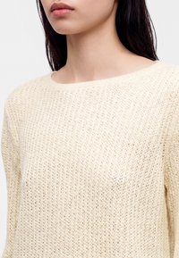 Femme portant un pull en tricot de couleur crème avec un col rond et de longs cheveux foncés visibles sur un fond clair.