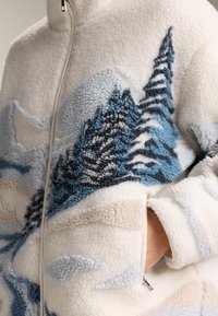 Jachetă crem din lână fleece cu model cu pini albaștri și gri, fermoar frontal și mână introdusă în buzunarul lateral.