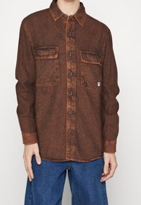 Brun denimskjorta med knappstängning framtill, två bröstfickor och krage. Har kontrasterande orange sömmar och knappar.