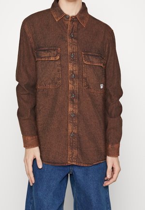 Chemise en denim marron avec des boutons sur le devant, deux poches poitrine et un col. Présente des coutures et des boutons orange contrastants.